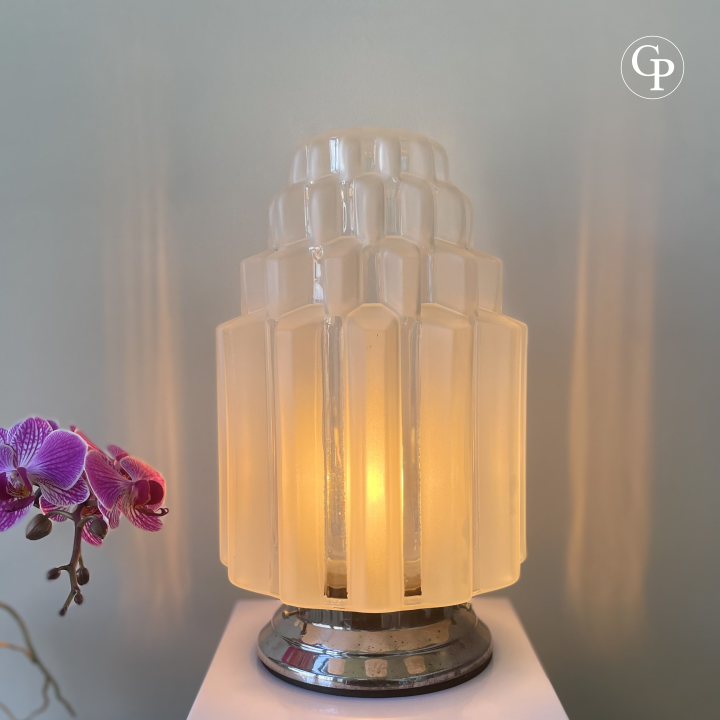 Vintage Art Deco table lamp glowing with warm light, featuring a tiered frosted glass shade and polished chrome base, displayed with purple orchids | Lampe de table vintage Art Déco allumée diffusant une lumière chaude, avec abat-jour en verre dépoli à étages et base chromée polie, mise en scène avec des orchidées pourpres