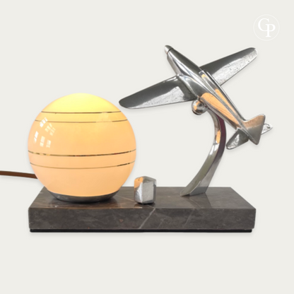 Lampe de table Art Déco vintage allumée avec une douce lumière chaude, mettant en valeur un avion Caudron Simoun chromé en position de vol dynamique à côté du globe opaline à rayures dorées sur base en marbre. Hommage à Antoine de Saint-Exupéry, vers 1937. | Vintage Art Deco table lamp illuminated with a warm glow, featuring a chrome Caudron Simoun airplane in dynamic flight position beside the lit opaline sphere with gold stripes on a marble base. Homage to Antoine de Saint-Exupéry, circa 1937.