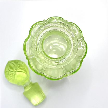 large and Top view of the rare Valambert n°12 Ouraline uranium glass perfume bottle showing its beautiful scalloped flower-shaped rim and the original stopper placed beside it, circa 1935. | Vue plongeante sur le flacon Valambert n°12 en verre à l'uranium Ouraline, mettant en valeur son élégant col festonné en forme de fleur et son bouchon d'origine posé à côté, vers 1935.