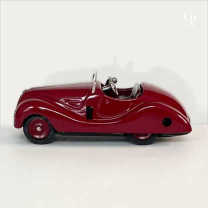 Full side profile view of the deep red Schuco Examico 4001 vintage tinplate roadster, highlighting the sleek aerodynamic design, windshield, and rubber tires on a light background. | Vue de profil complet de la voiture automate Schuco Examico 4001 couleur bordeaux, mettant en valeur sa silhouette aérodynamique, son pare-brise et ses pneus en caoutchouc sur fond clair.