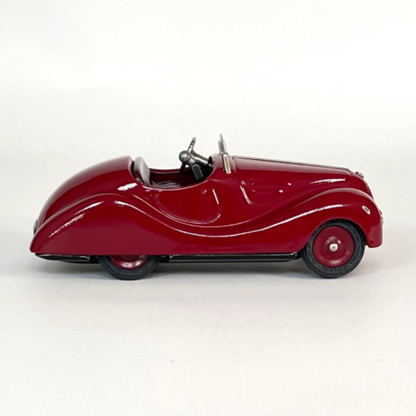 Right side profile view of the deep red Schuco Examico 4001 vintage tinplate roadster toy car, displaying its aerodynamic lines and black rubber tires on a light background. | Vue de profil droit de la voiture automate Schuco Examico 4001 de couleur bordeaux, montrant ses lignes aérodynamiques et ses pneus en caoutchouc noir sur fond clair.